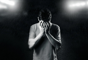 Un hombre de pie bajo la lluvia, con las manos en su rostro, mostrando una expresión de frustración o desesperación.